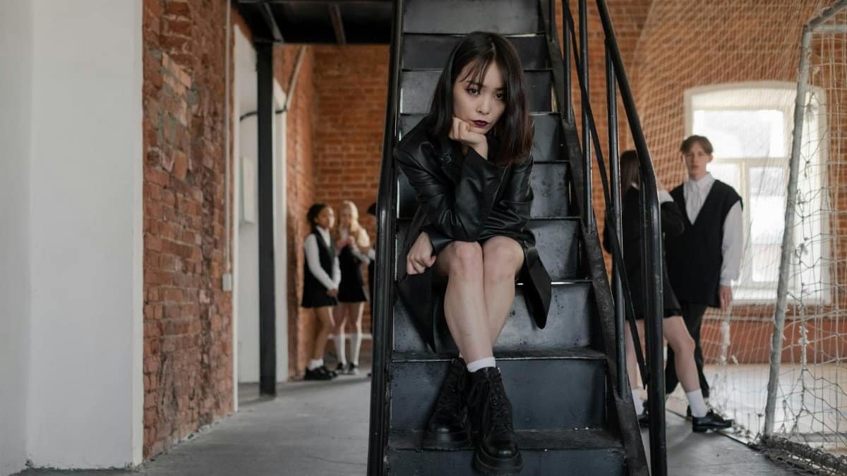 Teen girl sitting on stairs indoors with brick background, friends in distance.
