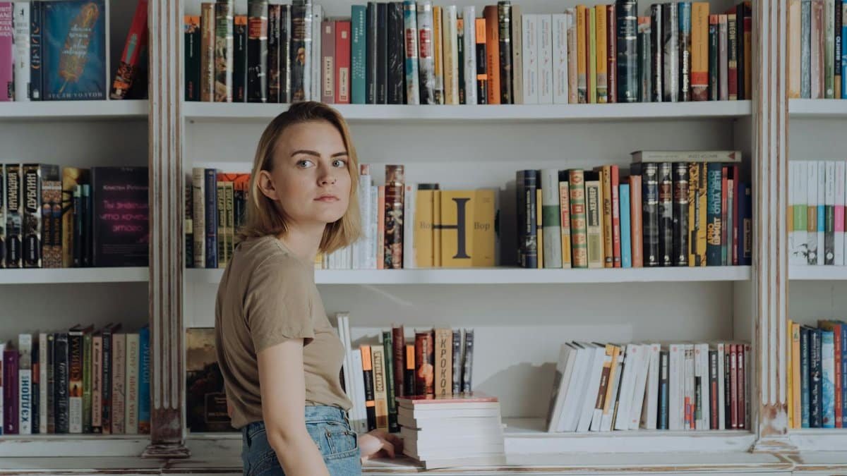 A young woman stands by bookshelves, surrounded by books, creating a serene library atmosphere.