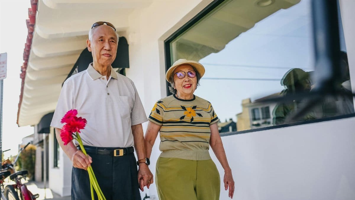 An elderly Asian couple walking hand-in-hand outdoors, enjoying a sunny day.