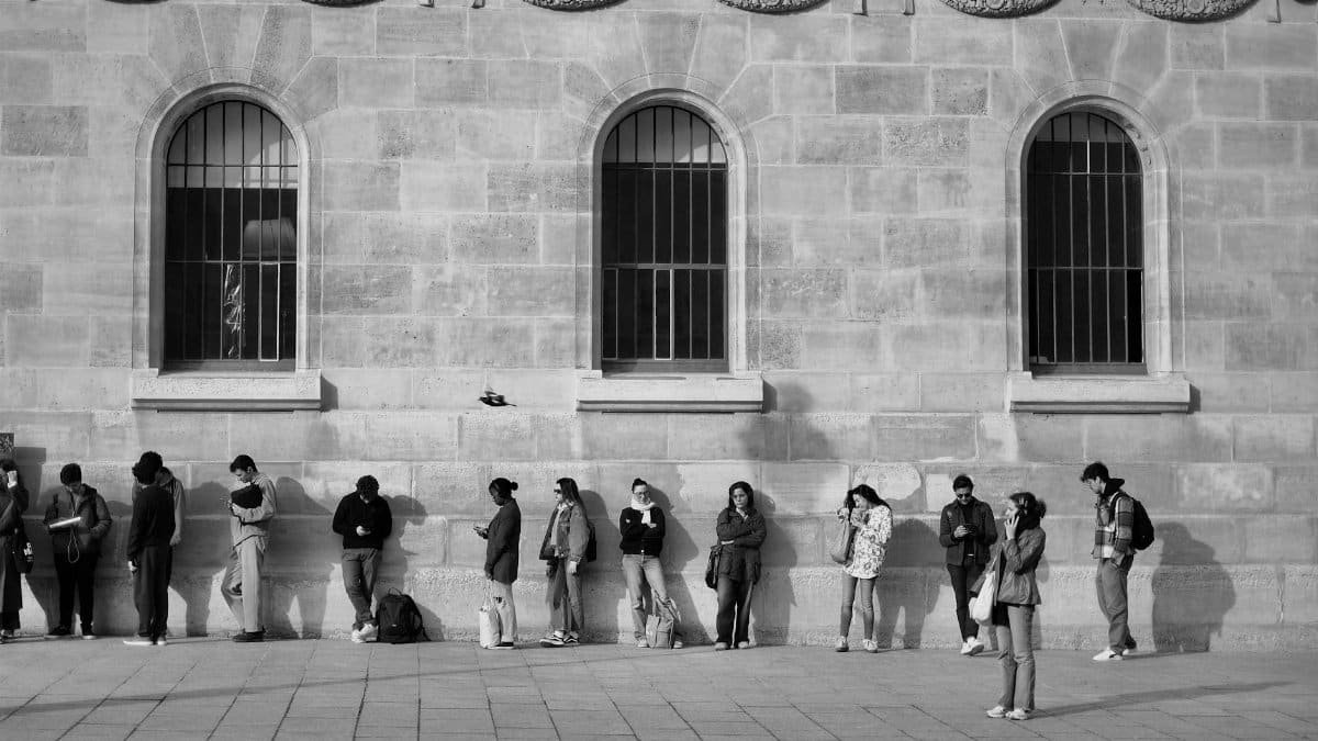 Black and white photo showing a group of people waiting outside a historic stone building with arched windows.
