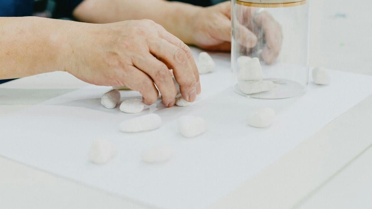 Hands arranging white stones on paper with a glass jar, highlighting creativity and art.