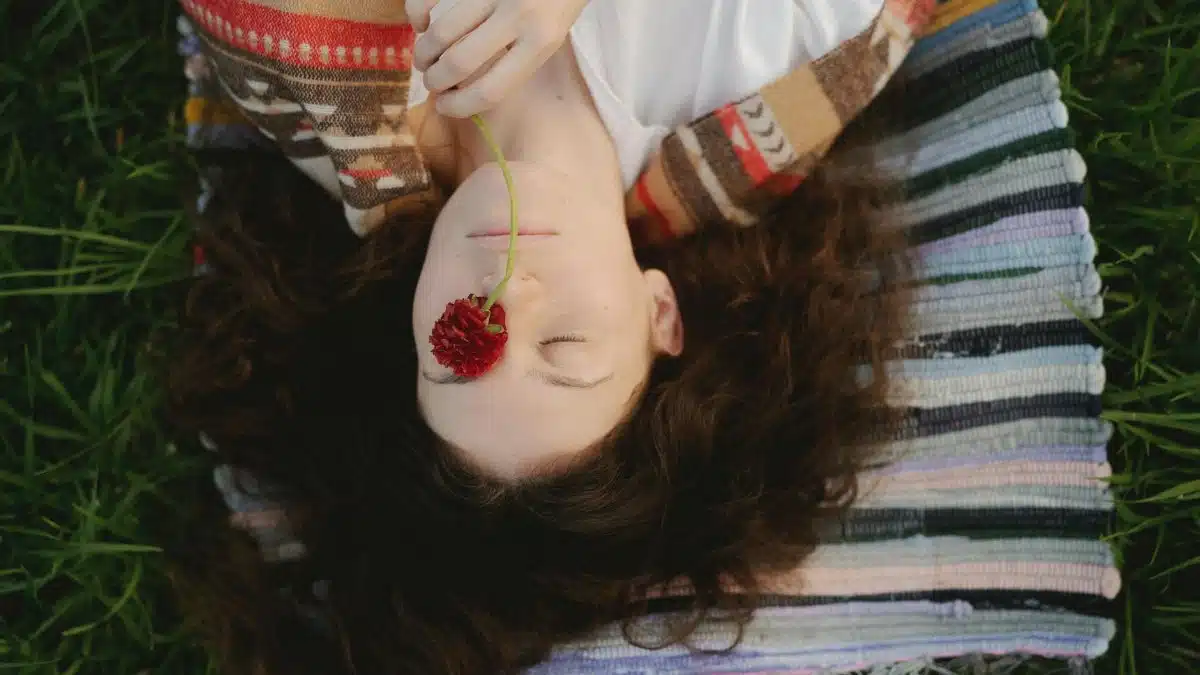 A woman lies on a colorful rug outside, holding a red flower, exuding peace and tranquility.