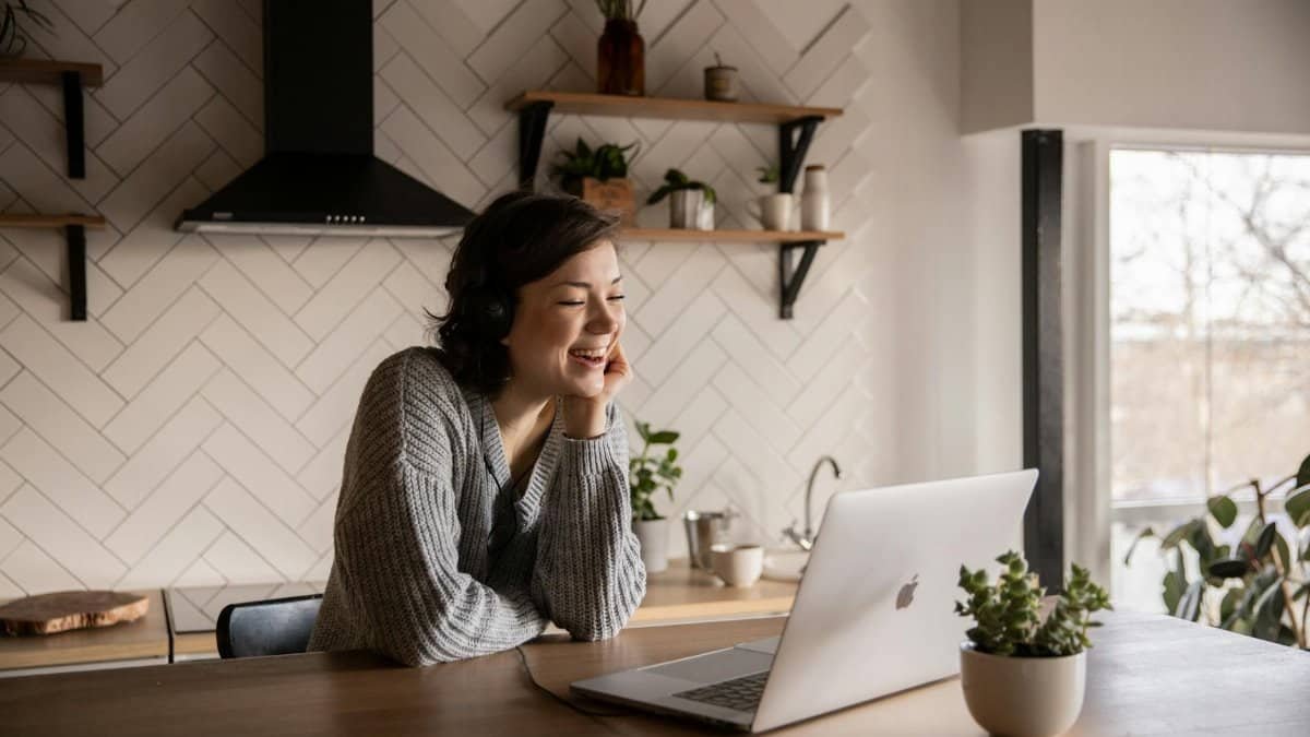 Young cheerful female smiling and talking via laptop while sitting at wooden table in cozy kitchen
