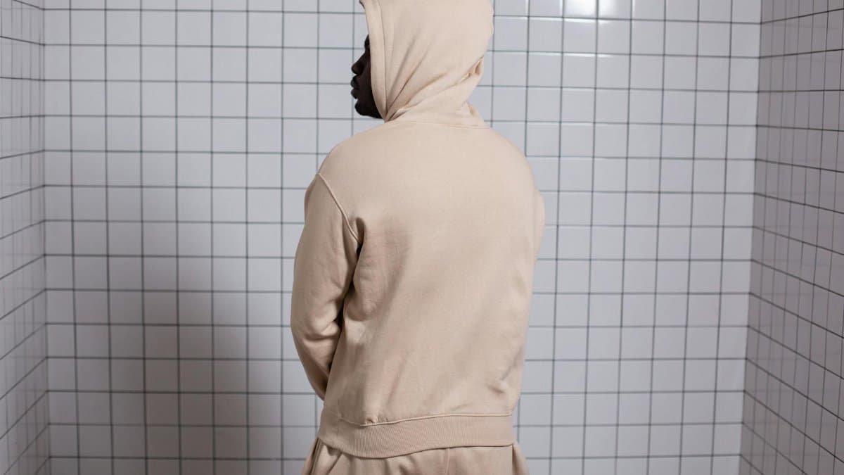 A man in a beige hoodie stands thoughtfully against a tiled wall, creating a calm and modern scene.
