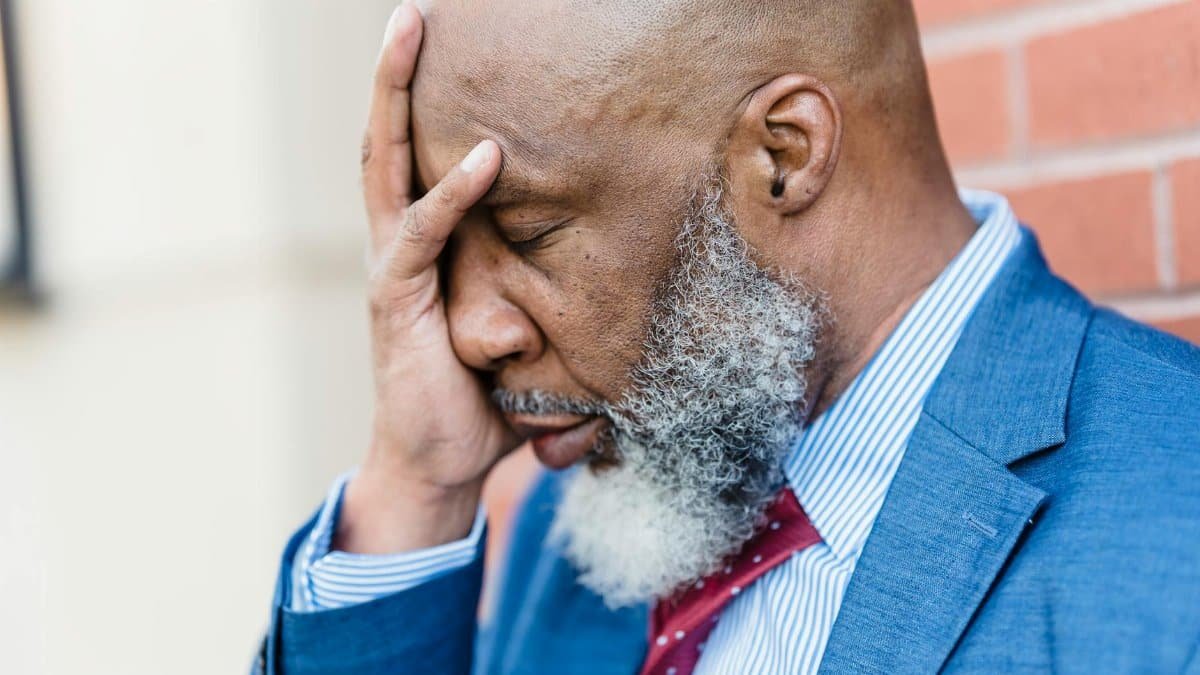 A middle-aged man in a blue suit shows stress and despair with hand on face.