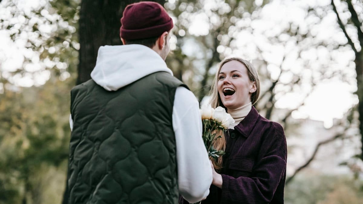 Loving boyfriend in hat giving bunch of roses as gift to happy girlfriend with long hair in casual warm clothes in park