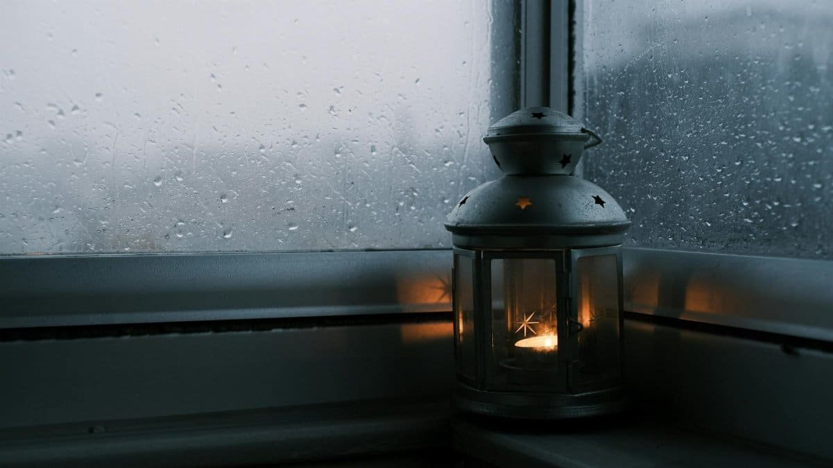 A flickering lantern by a rain-streaked window creates a cozy ambiance.