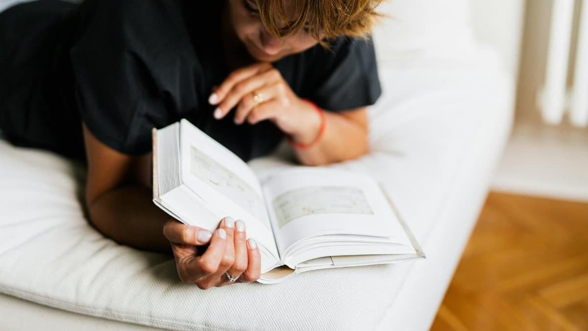 A woman lying comfortably on a bed, engrossed in reading a book at home, evokes a sense of relaxation and leisure.