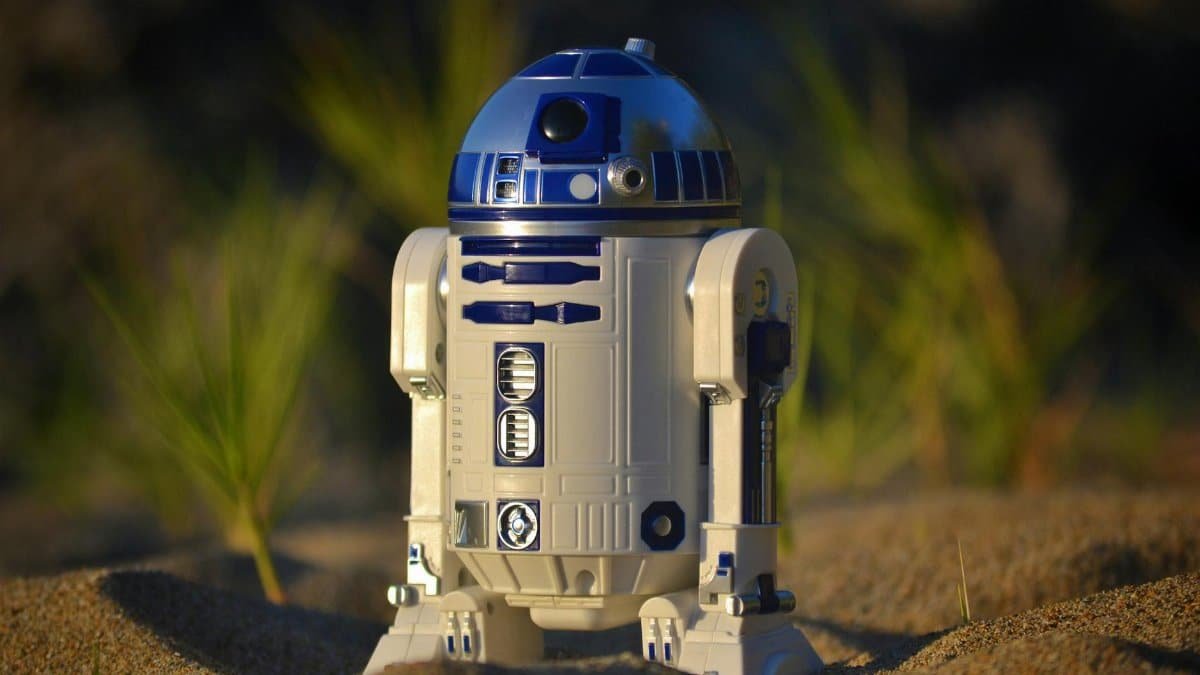 Close-up of an R2-D2 toy standing on sand, set against a green background in bright daylight.