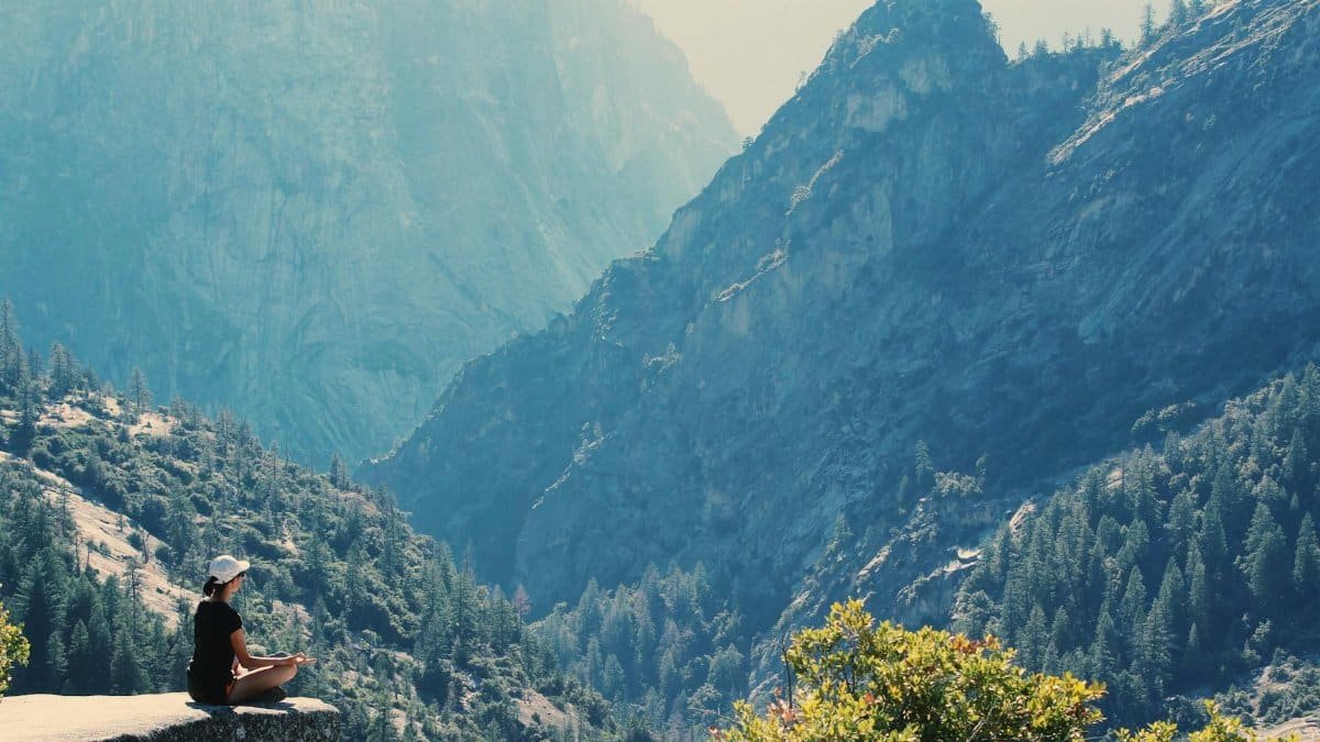 A woman practices meditation on a cliff edge, surrounded by majestic mountains and serene nature.