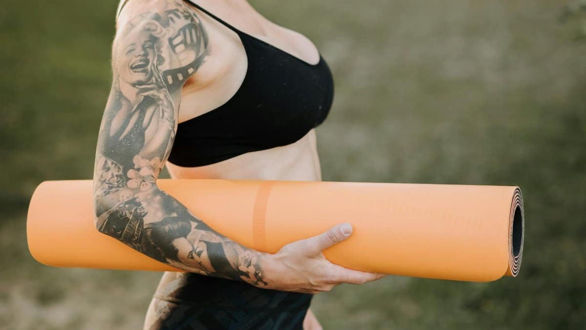 Side view of crop anonymous tattooed female in sports clothes with rolled yoga mat standing on lawn