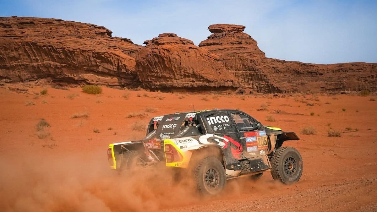 Off-road racing car speeds through red desert terrain with dramatic rock formations.