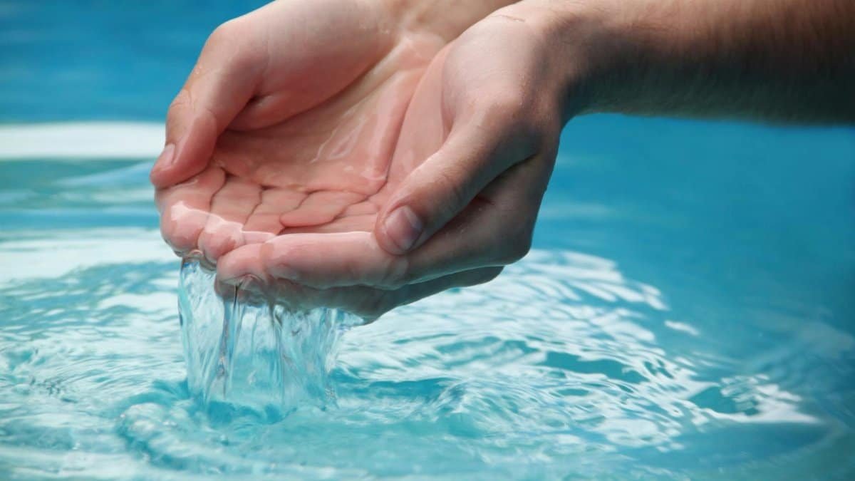 Close-up of hands scooping clear blue water from a reflective pool outdoors.