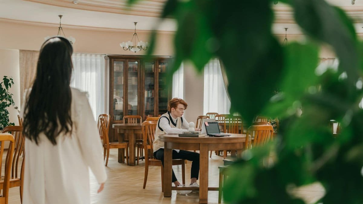 A woman approaches a man working on a laptop in a library setting.
