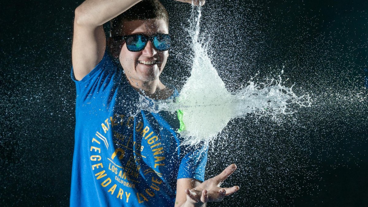 Man in sunglasses joyfully bursts a water balloon, creating dynamic splash in dark setting.