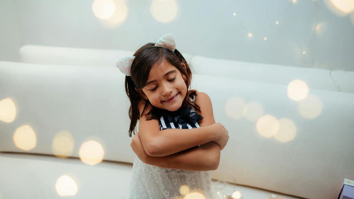 Cute child wearing cat ears, hugging herself amidst glowing fairy lights indoors.
