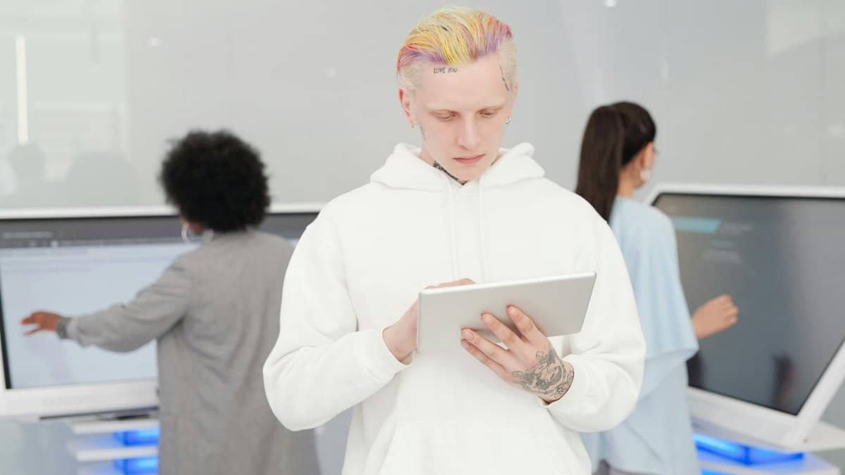 A young man with colorful hair and tattoos using a tablet in a modern tech environment while others work in the background.