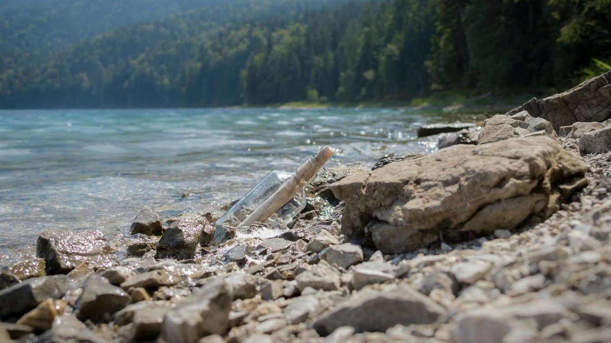 A glass bottle with a message rests on a rocky lakeside shore, surrounded by nature and tranquil waters.