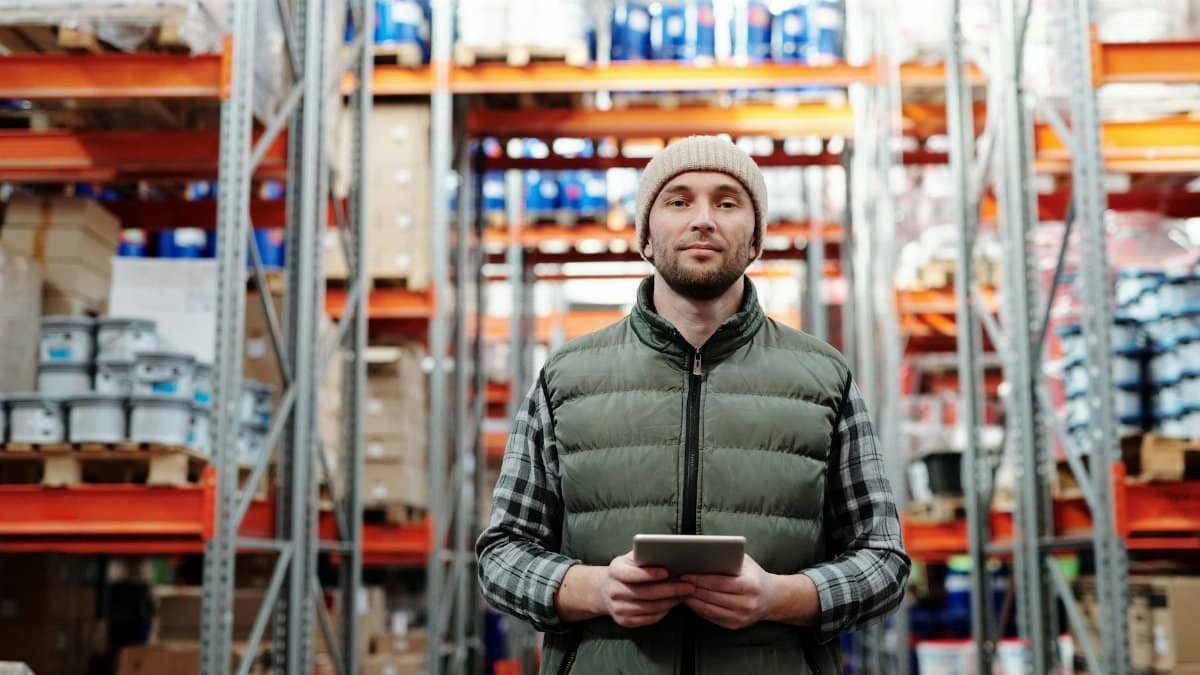 A warehouse worker wearing a beanie uses a tablet to manage inventory in a storeroom with shelves.