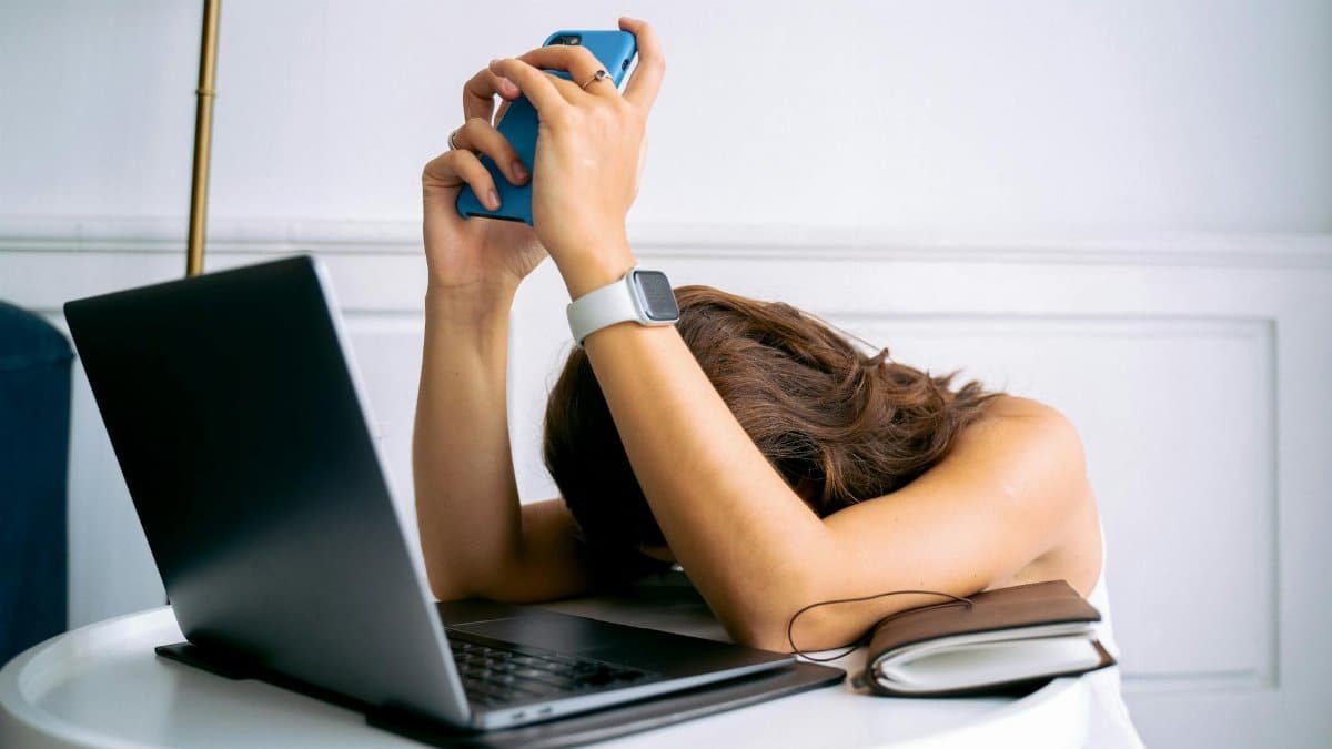 A woman overwhelmed by work, resting head on table with laptop, phone, and smartwatch.