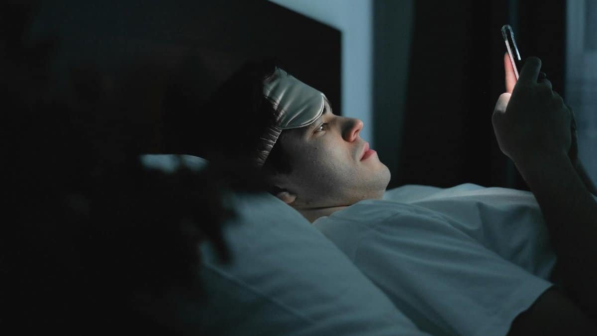 A man wearing a sleep mask late at night using a smartphone on his bed.