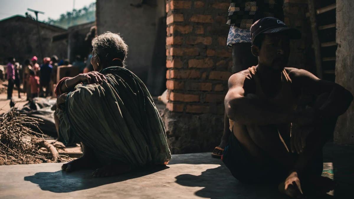 A group of adults sitting outdoors in a village setting, casting strong shadows on a sunny day.