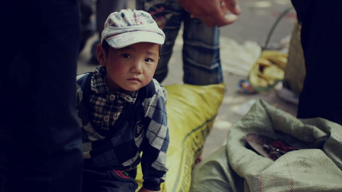 A young child wearing a cap gazes thoughtfully amidst an outdoor market setting, surrounded by sacks.