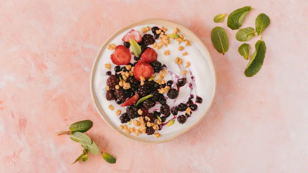 Top view of a vibrant yogurt bowl with mixed berries, granola, and mint leaves on a pink background.