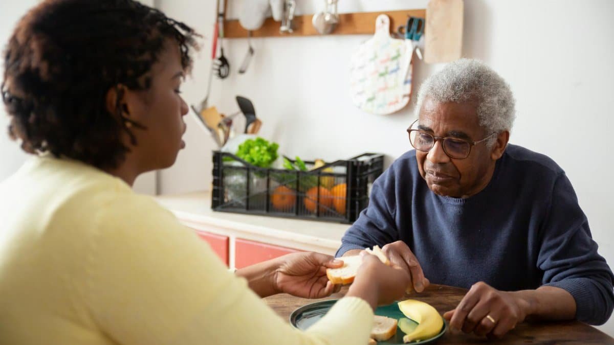 A woman assists an elderly man with meals at home, showcasing care and support.