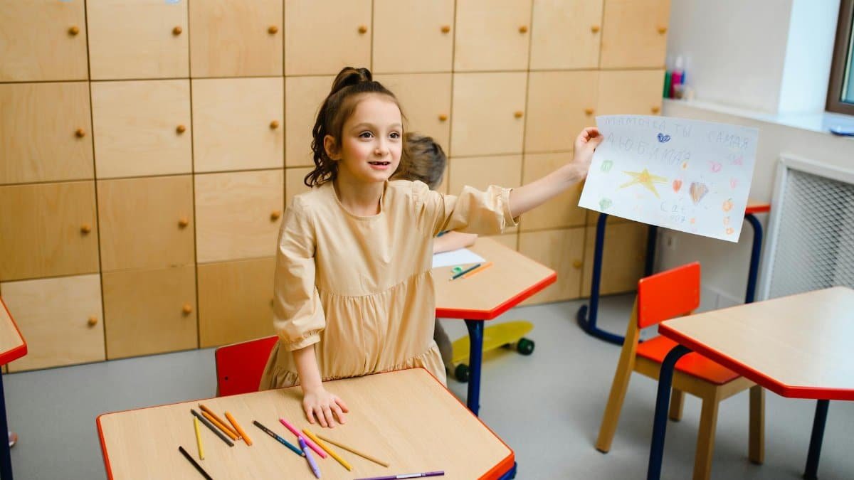 Smiling girl proudly showing her drawing in a vibrant kindergarten classroom.