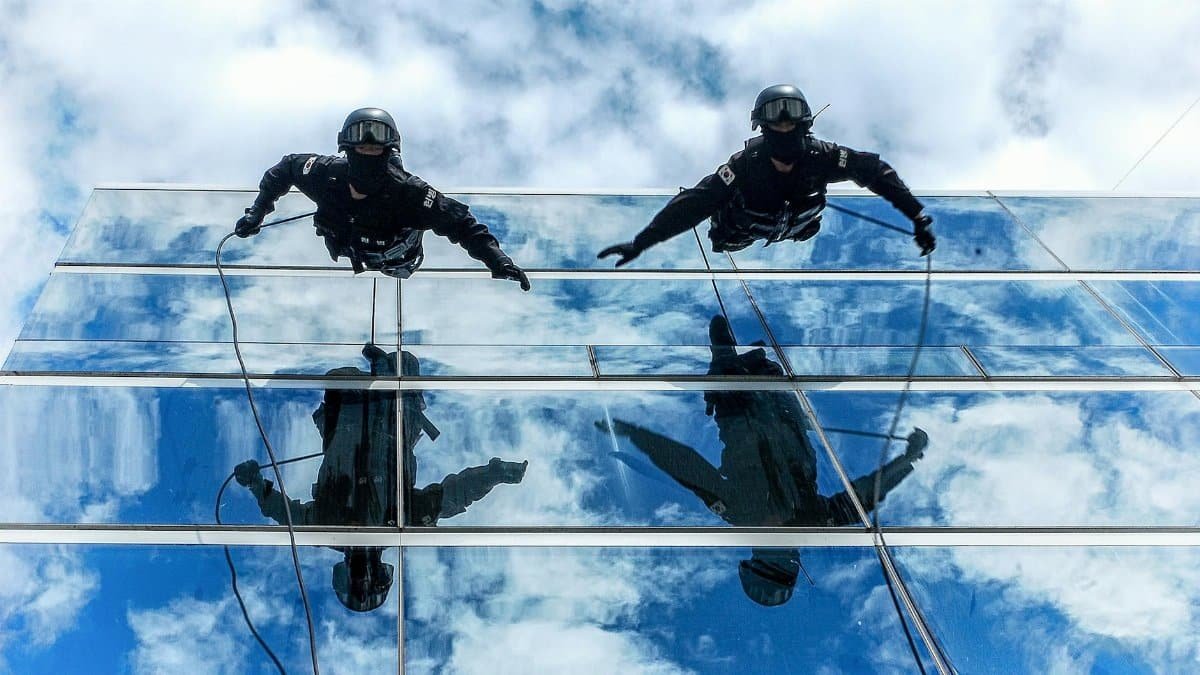 Gendarmerie officers rappelling down a glass building with sky reflections.