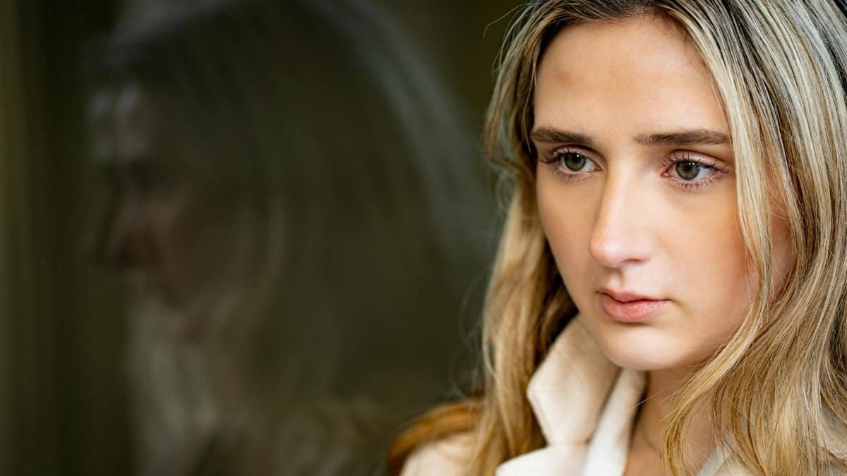 Portrait of a young woman with blonde hair gazing out a window with a reflection.