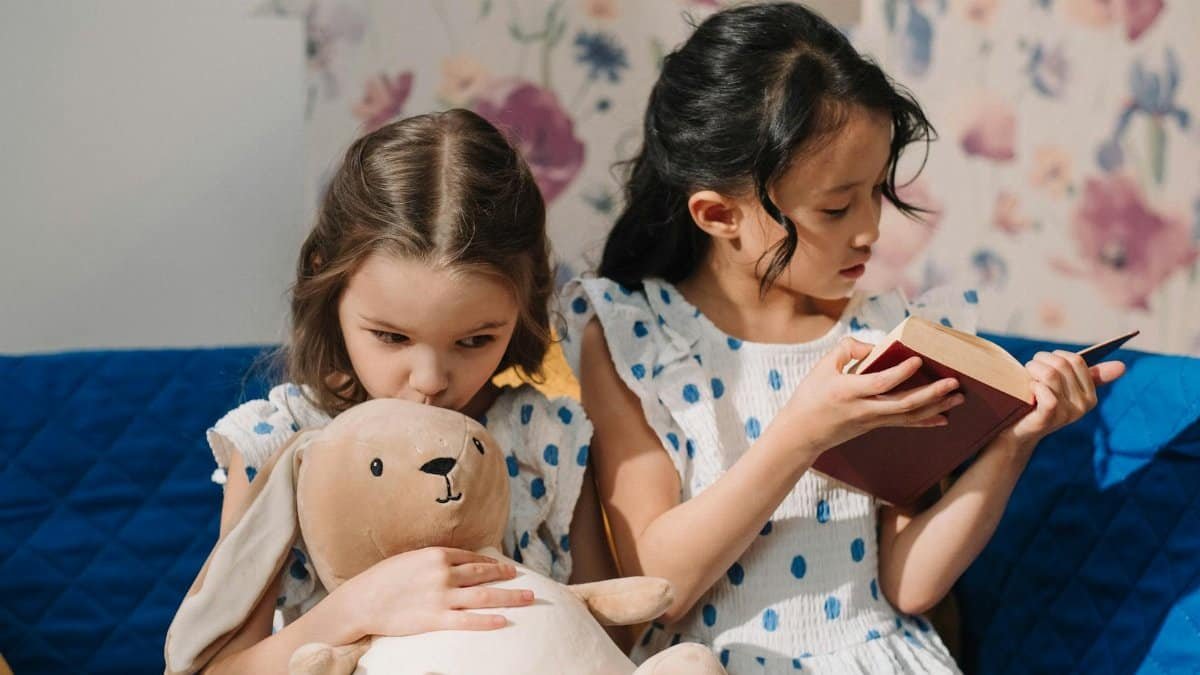 Two young girls in white dresses reading and cuddling a plush toy on a bed.