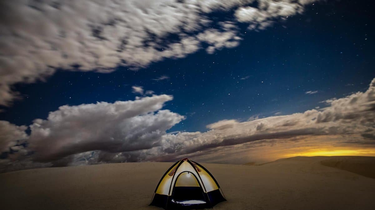A solitary tent under a starry night in a vast desert, with dramatic skies and distant sunset glow.