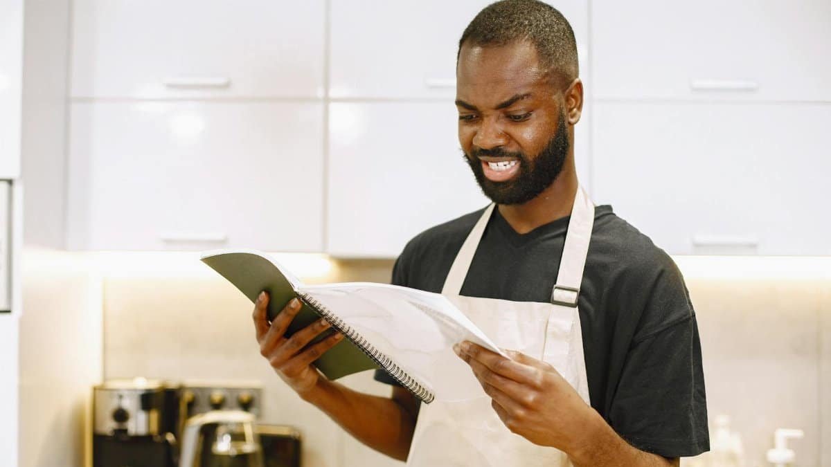 A chef wearing an apron looks puzzled while reading a recipe in a contemporary kitchen.