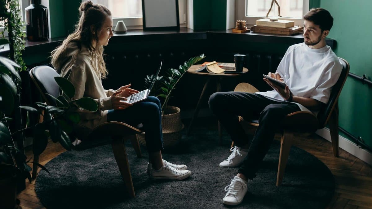 A man and woman engaged in conversation at a stylish, plant-filled home office with modern furniture and digital devices.