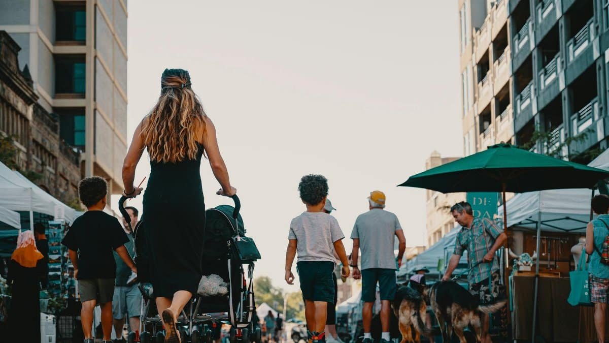Family walking with stroller and dogs through lively outdoor market street on a sunny day.