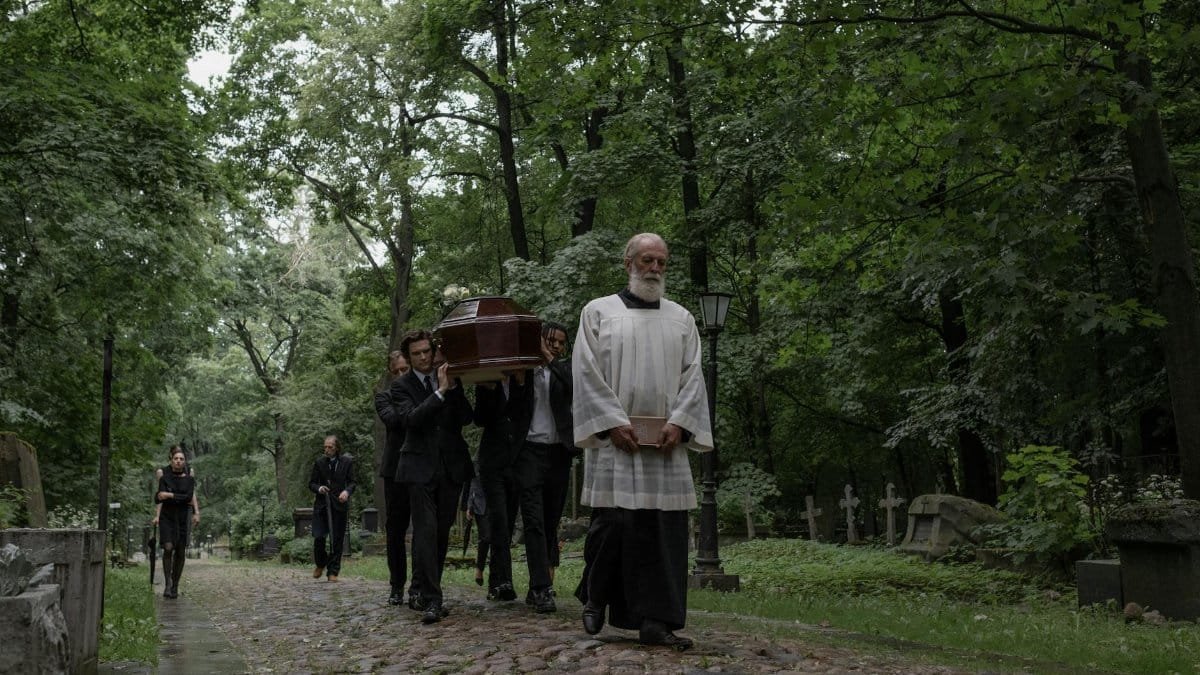 A group of mourners carrying a casket through a tranquil, tree-lined cemetery.