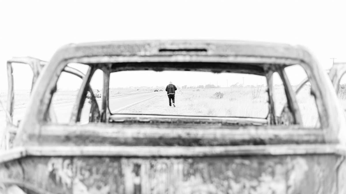 Artistic black and white photo capturing a solitary figure through the frame of an abandoned car.