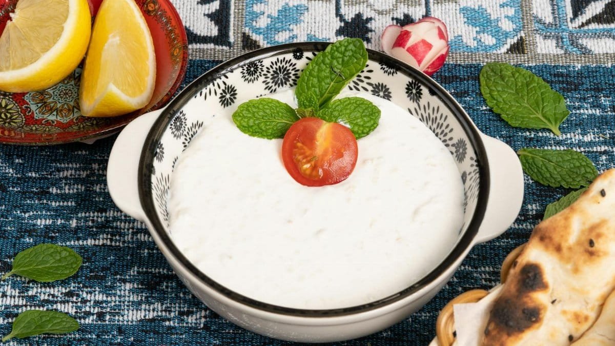 Delicious bowl of creamy yogurt garnished with mint leaves and a cherry tomato, surrounded by fresh bread and lemon slices.