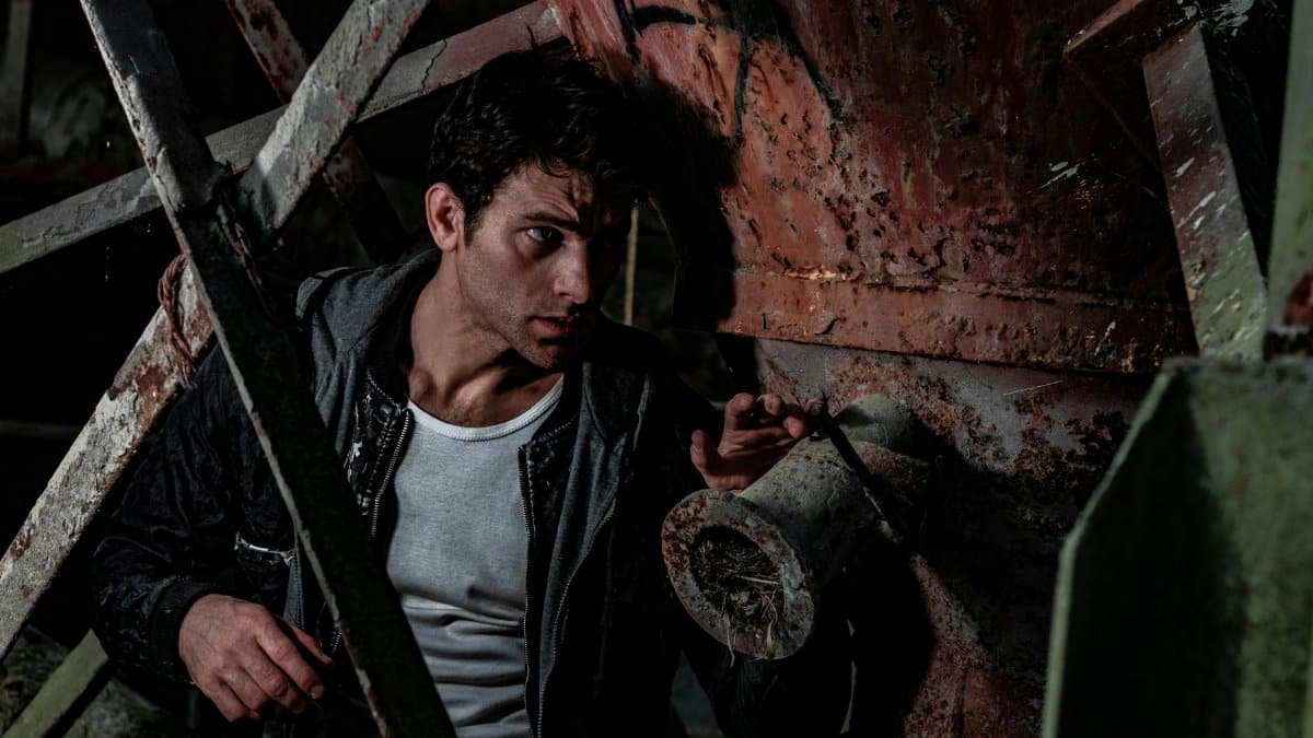 Man hidden in a dark industrial setting, appearing cautious and tense in a rusty environment.