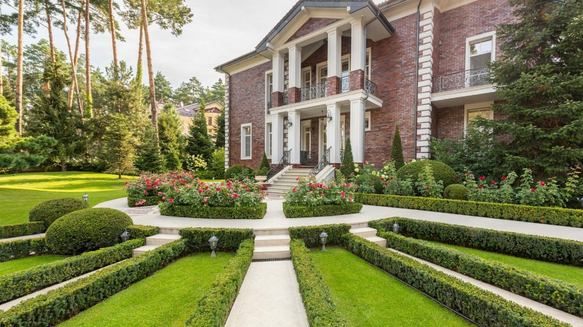 Luxurious brick mansion surrounded by lush gardens and trimmed hedges on a sunny day.