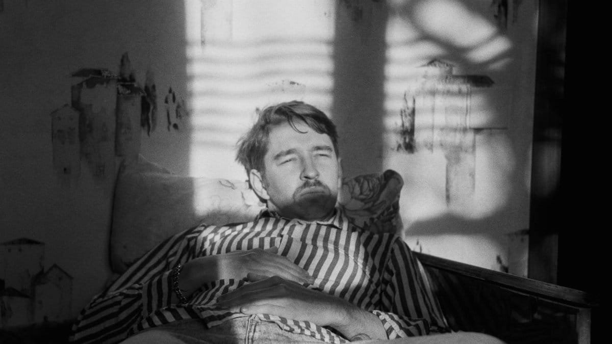 A contemplative man lounges on a chair in soft window light, casting intricate shadows.