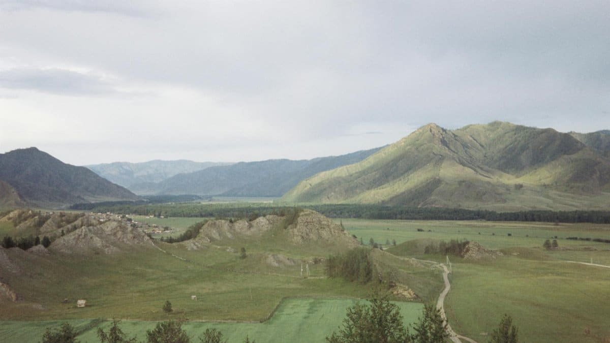 A breathtaking view of a mountain valley with lush green fields and a distant village under a cloudy sky.