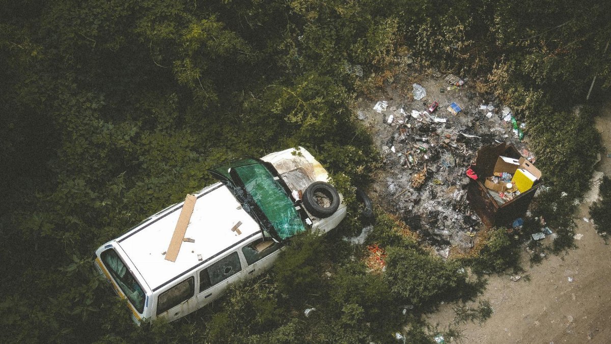 Overhead view of a damaged car and trash in a forested area indicating pollution and neglect.