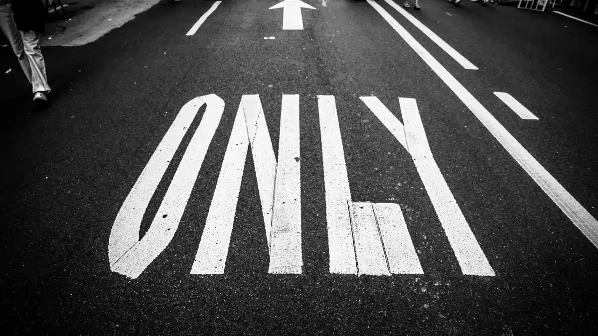 Black and white photo of road, showing traffic markings, a crowd, and an arrow.