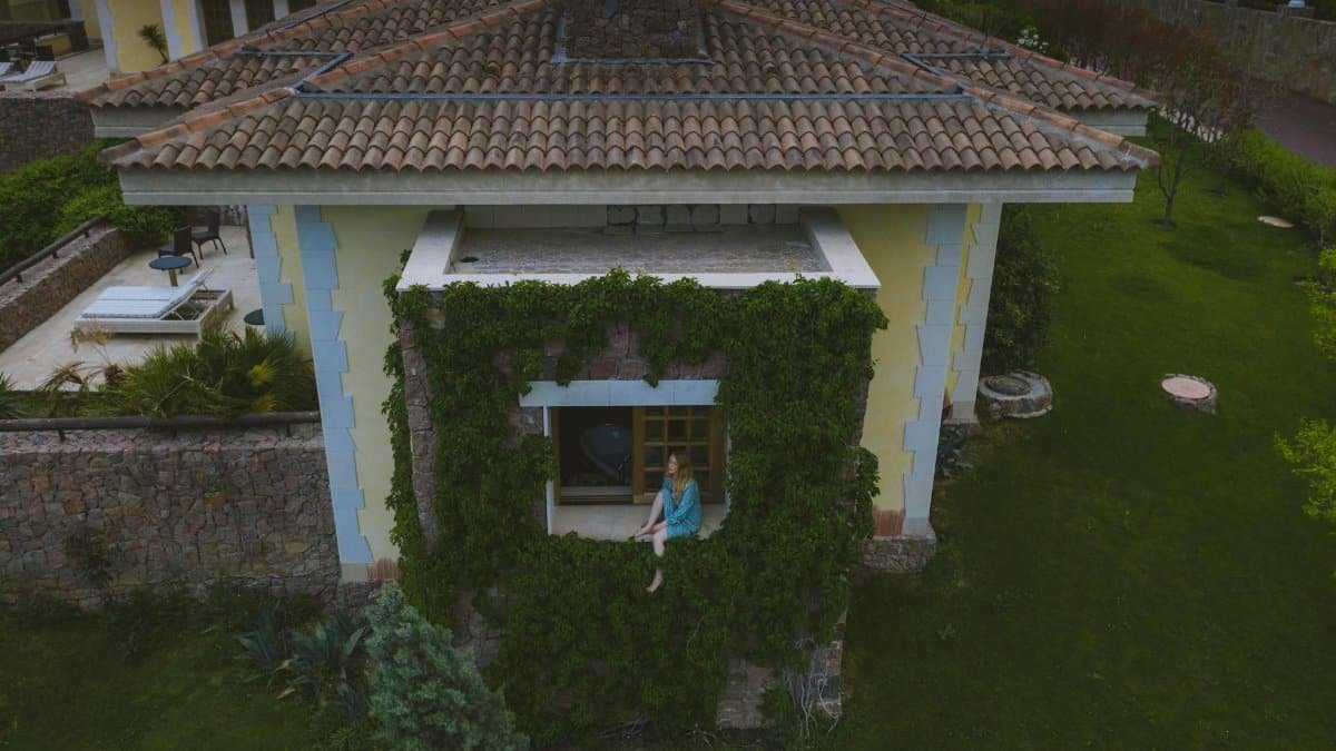 Aerial view of a woman sitting on a window sill of a vine-covered house in a garden setting.
