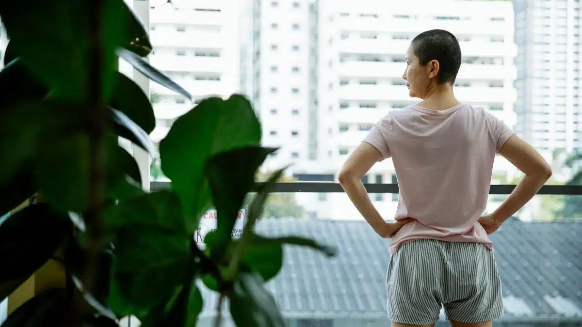 Back view of ethnic ill female with hands on waist looking away on balcony of hospital