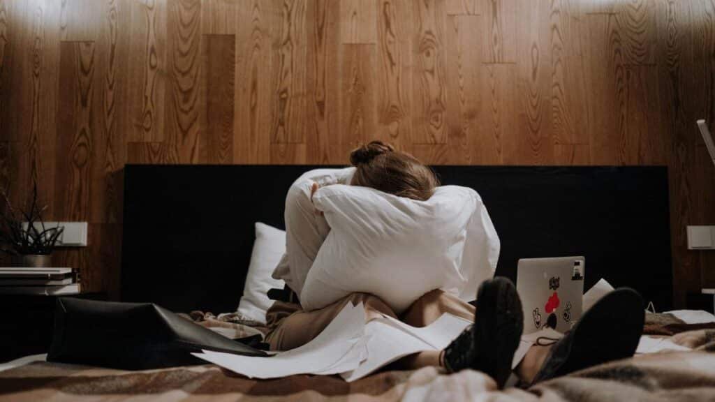 A woman sits on a bed clutching a pillow, surrounded by papers and a laptop, conveying a sense of stress.