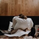 A woman sits on a bed clutching a pillow, surrounded by papers and a laptop, conveying a sense of stress.